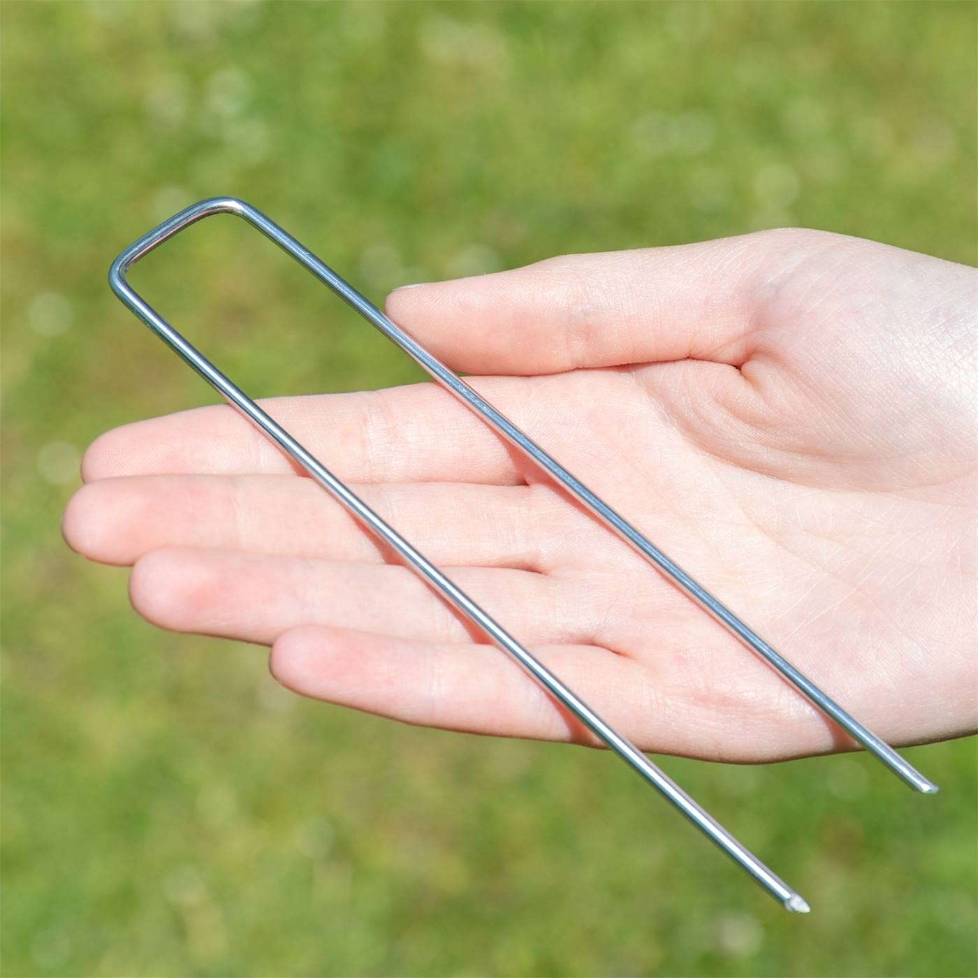 En hand håller ett VOSS.farming U-format markankare (150x150x25 mm, Ø2,7 mm) för fleece, staket och nät mot en suddig gräsbakgrund - ingår i en sats om 50 delar.