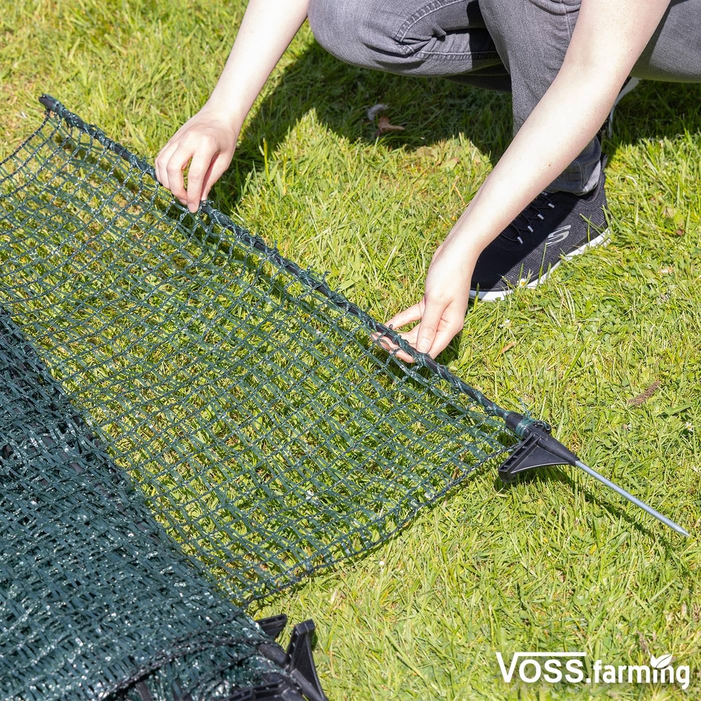 Dubbelklicka för att förstora En person ställer upp VOSS.farming classic 20 m stängsel, ett 80 cm högt grönt trådstängsel med 12 stolpar, på gräs. VOSS.farming-logotypen finns i det nedre högra hörnet.