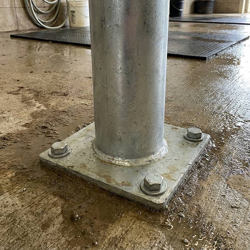 Growi®-fäststolpen på 170 cm med rundrör och bottenplatta (Ø 102 mm) förankras med fyra stora bultar i ett vått betonggolv; i bakgrunden syns vatten och en slang.
